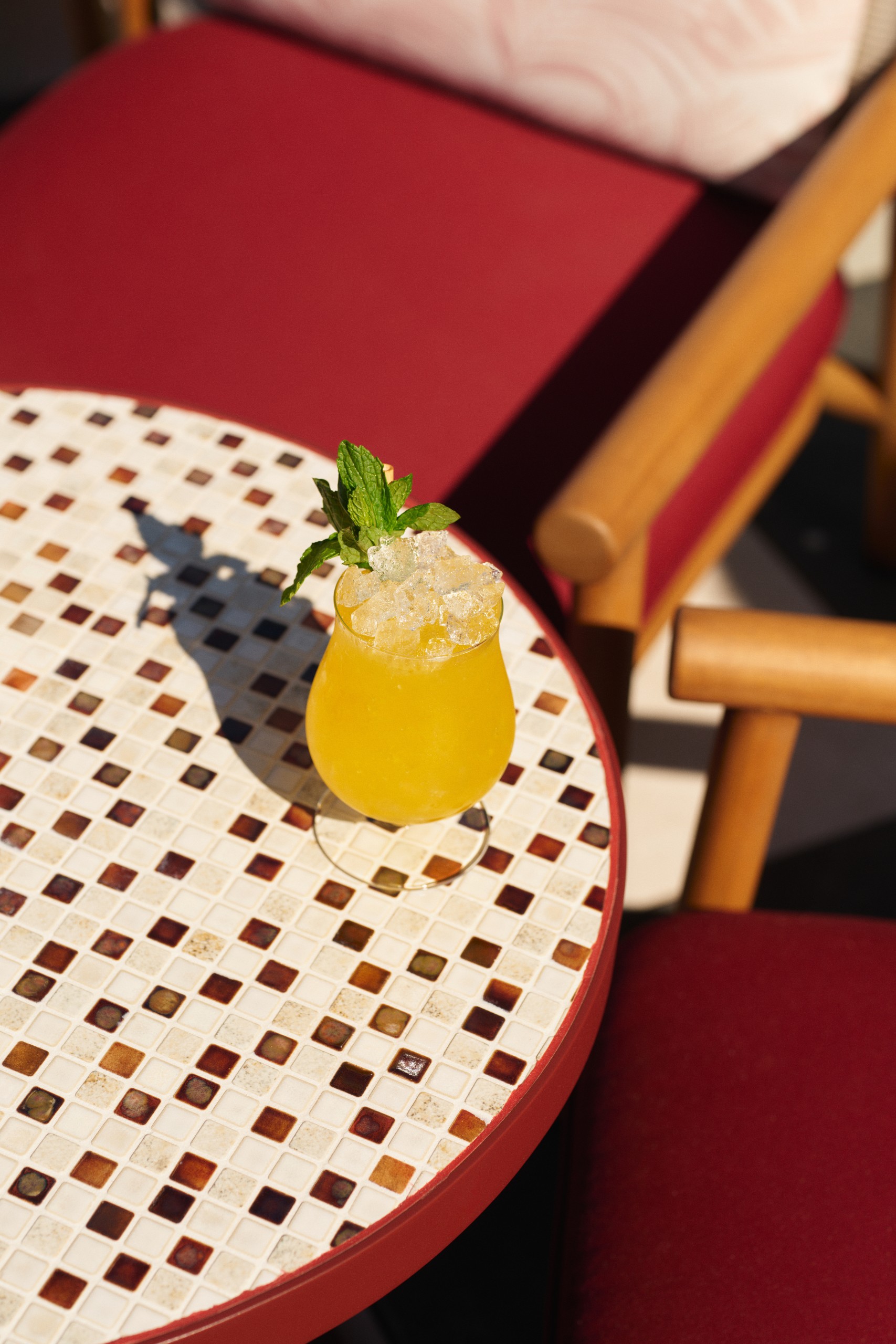A refreshing yellow cocktail with crushed ice and mint garnish sits on a patterned mosaic table beside a stylish lamp and red chair.