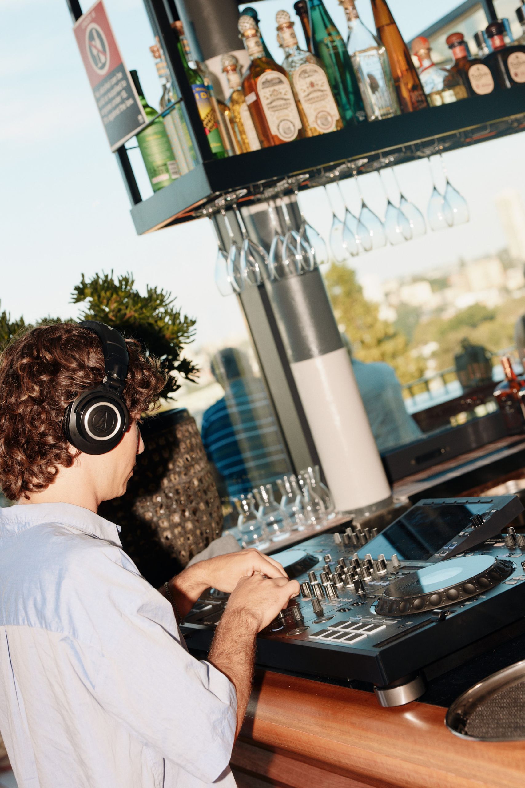 A DJ wearing headphones mixes music on a rooftop, surrounded by bottles and wine glasses against a cityscape backdrop under a clear sky.